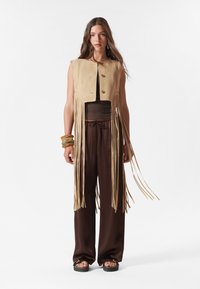 Beige suede cropped vest with fringe detailing, paired with brown loose-fitting pants. Notable accessories include wooden bangles and platform sandals.