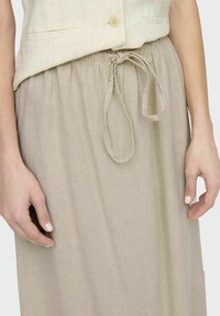 Falda de lino beige con cintura fruncida y lazo de cordón. Textura suave y caída ligera. Dos manos sosteniendo suavemente la tela.