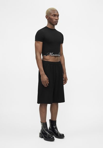 Άνδρας με κοντά ξανθά μαλλιά στέκεται φορώντας μαύρο κοντό μπλουζάκι με κείμενο, μαύρα φαρδιά σορτς και μαύρα γυαλιστερά αθλητικά μποτάκια με φερμουάρ.