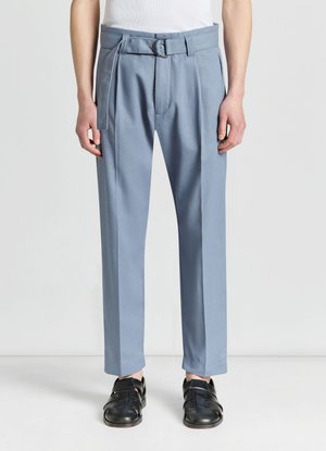 Uomo che indossa pantaloni su misura azzurri con cintura abbinata e sandali neri chiusi in punta, in piedi su un pavimento bianco.