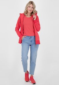 Woman standing with one hand in pocket and the other holding her red zip-up hoodie, wearing a red striped shirt, blue jeans, and red sneakers.