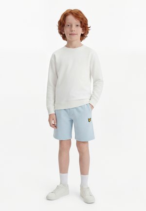 Copil cu păr roșu, stând orientat spre înainte, îmbrăcat într-un tricou alb cu mâneci lungi, pantaloni scurți bleu deschis cu logo galben, șosete albe și adidași albi.