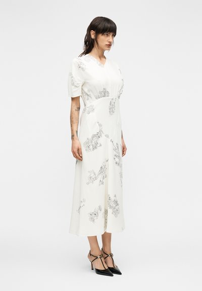 Λευκό μεσαίο φόρεμα με λουλουδένια σχέδια γ ραμμής, κοντά μανίκια με φουσκάλες, λαιμόκοψη σε σχήμα V, εφαρμοστή μέση και φούστα με καμπάνα. Φορεμένο με μαύρα μυτερά τακούνια.