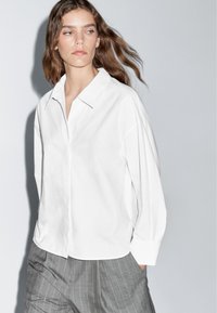 Hvid skjorte med knapper og krave, i en løs pasform, kombineret med grå stribede bukser. Stoffet ser glat ud med et minimalistisk design.