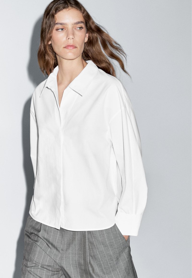 Hvid skjorte med knapper og krave, i en løs pasform, kombineret med grå stribede bukser. Stoffet ser glat ud med et minimalistisk design.