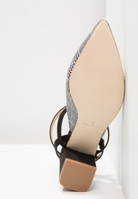 Chaussure à talon pour femme à bout pointu avec dessus en tissu à carreaux, brides de cheville noires et talon bloc, semelle tournée vers l'avant sur fond blanc.