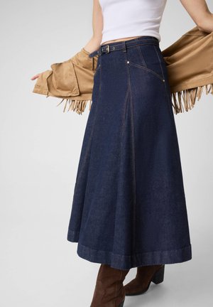 Femme portant une longue jupe en jean bleu foncé avec une ceinture, des bottes marron, un haut blanc, et tenant ouverte une veste fauve à franges derrière elle.