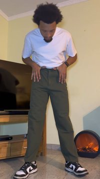 Casual outfit featuring a white t-shirt and dark green trousers. The person wears black and white sneakers. Background includes a TV and a fireplace.