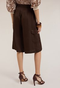 Culotte di seta marrone con vita elasticizzata e tasche laterali, abbinate a sandali con tacco borgogna con un design a tracolla.