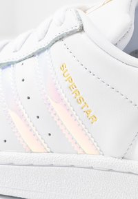 Bílá kožená teniska se zlatým nápisem "SUPERSTAR". Má duhové tři pruhy a strukturovanou bílou gumovou podrážku. Zaoblený design špičky.