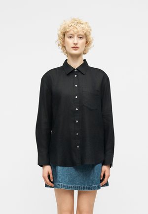 Blond, krullend persoon met een zwart geruit overhemd en een blauwe spijkerrok, staand tegen een effen witte achtergrond.