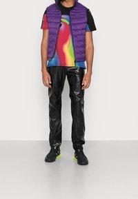 Lila pufferväst över en färgglad abstrakt t-shirt, i kombination med svarta glansiga byxor och svarta sneakers med gröna accenter.