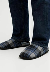 Chaussons à carreaux en laine grise, bleue et noire avec une texture douce ; une semelle en caoutchouc noire ; associés à un pantalon en denim foncé.
