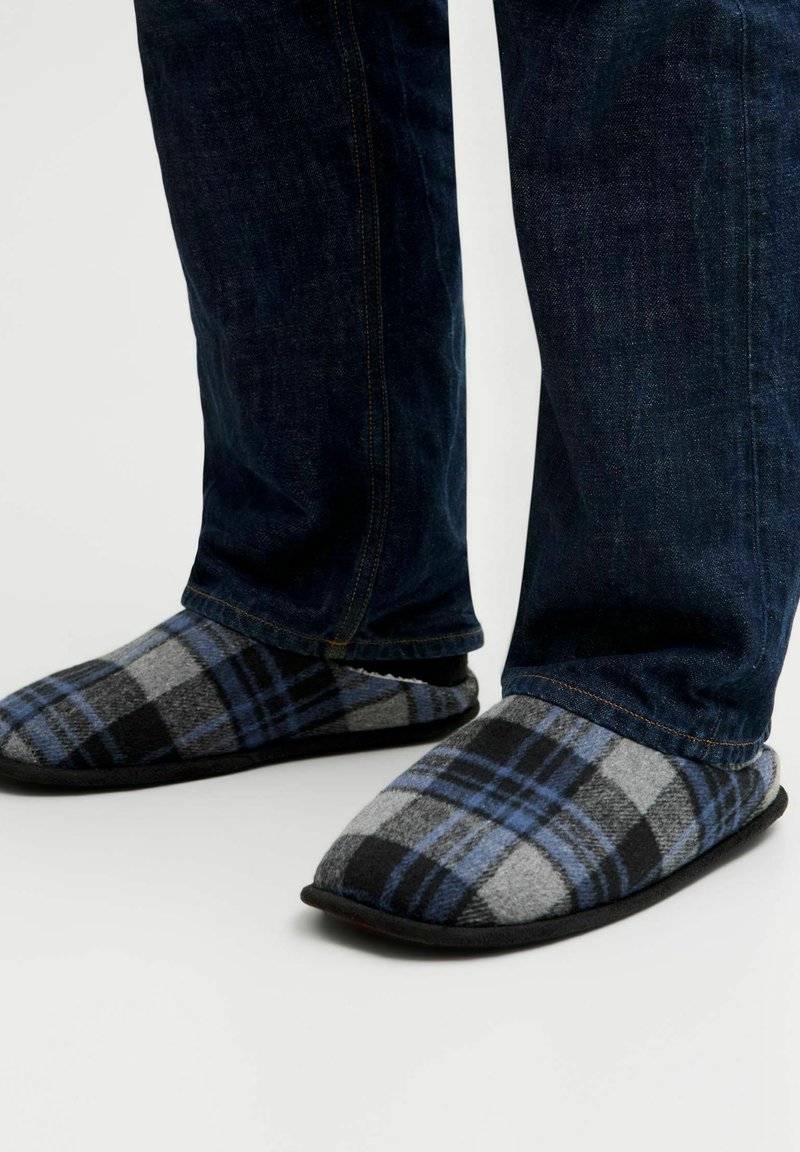 Chaussons à carreaux en laine grise, bleue et noire avec une texture douce ; une semelle en caoutchouc noire ; associés à un pantalon en denim foncé.