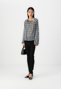Top trasparente a scacchi bianco e nero con maniche lunghe e scollatura arricciata, abbinato a pantaloni neri slim-fit e scarpe nere a punta.