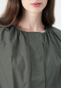 Blouse vert olive avec un col rond et un tissu froncé. Comprend des coutures horizontales et une patte de boutonnage à l'avant. Texture légère et lisse.