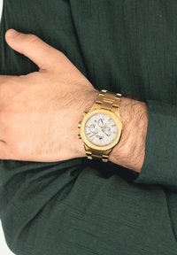 Guldtonet armbåndsur med en struktureret sølvskive, tre underdials og et poleret metalarmbånd, båret på en hånd mod en mørkegrøn baggrund.