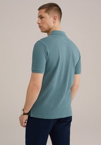 Teal spalvos polo marškiniai iš trumpomis rankovėmis, su klasikiniu apykaklės dizainu ir tiesiu kraštu. Dėvimi su tamsiai mėlynomis kelnėmis ir rudomis laikrodžiu.