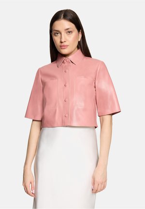 Femme aux longs cheveux foncés portant une chemise en cuir rose courte à boutons et une jupe blanche taille haute, debout devant un fond uni.