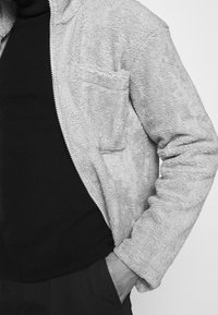 Lys grålæder fleece zipjakke med tekstureret overflade, frontlomme og ribbede manchetter, lagd oven på en sort top.