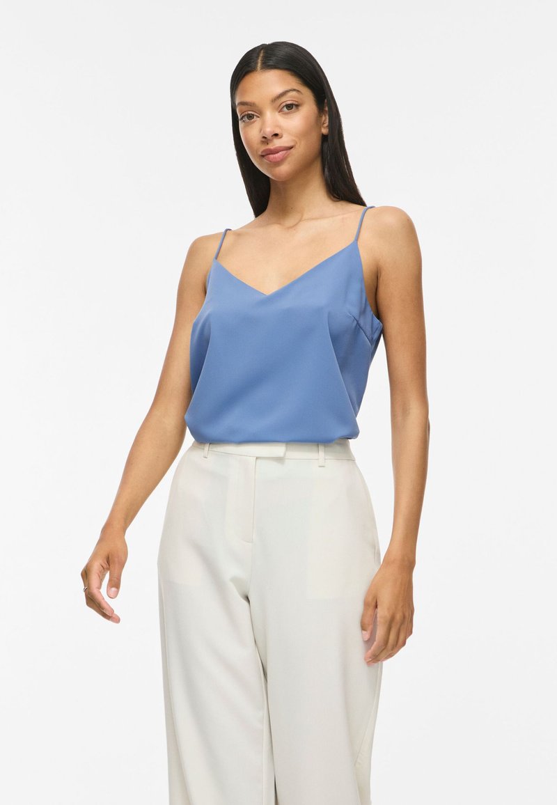 Femme aux longs cheveux foncés portant un haut bleu sans manches et un pantalon blanc taille haute, debout devant un fond blanc uni.