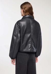 Persona con capelli castani che indossa una giacca in pelle nera oversize e pantaloni neri, in piedi di spalle contro uno sfondo chiaro.