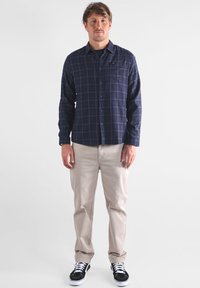 Homme se tenant de face, portant une chemise à carreaux bleu marine, un pantalon beige et des baskets noires avec des lacets blancs sur un fond blanc.