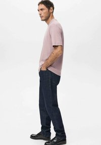 Light pink short-sleeve t-shirt, medium-fit, paired with dark blue straight-leg jeans. Model stands with hands in pockets, wearing black shoes.