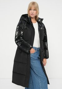 Zwarte gewatteerde jas met een glanzende, glimmende afwerking, capuchon en voorzakken. Heeft een lange lengte en is gecombineerd met lichtblauwe jeans.