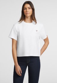 Weißes Baumwoll-Cropped-T-Shirt mit kurzen Ärmeln, rundem Halsausschnitt und einem kleinen dreieckigen Logo auf der Brust. Kombiniert mit dunklen Jeans.