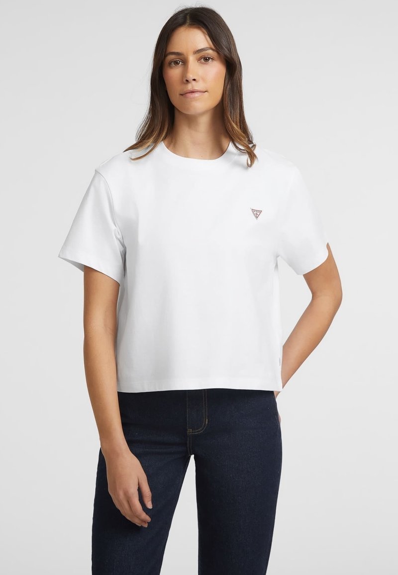Weißes Baumwoll-Cropped-T-Shirt mit kurzen Ärmeln, rundem Halsausschnitt und einem kleinen dreieckigen Logo auf der Brust. Kombiniert mit dunklen Jeans.