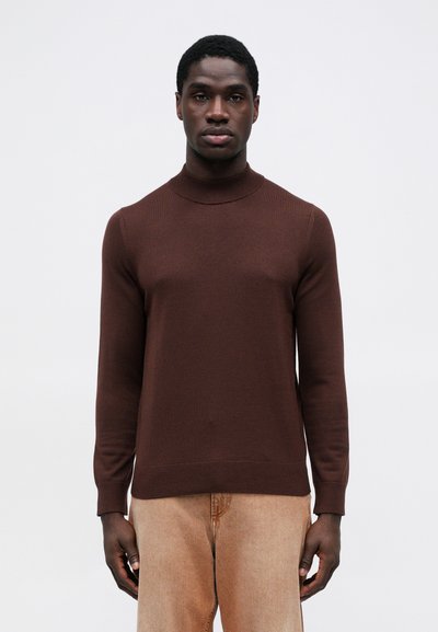 Smeđi, krojeni turtleneck pulover s dugim rukavima, od glatkog pletenog materijala. Uparen s svijetlosmešim hlačama za kontrast.