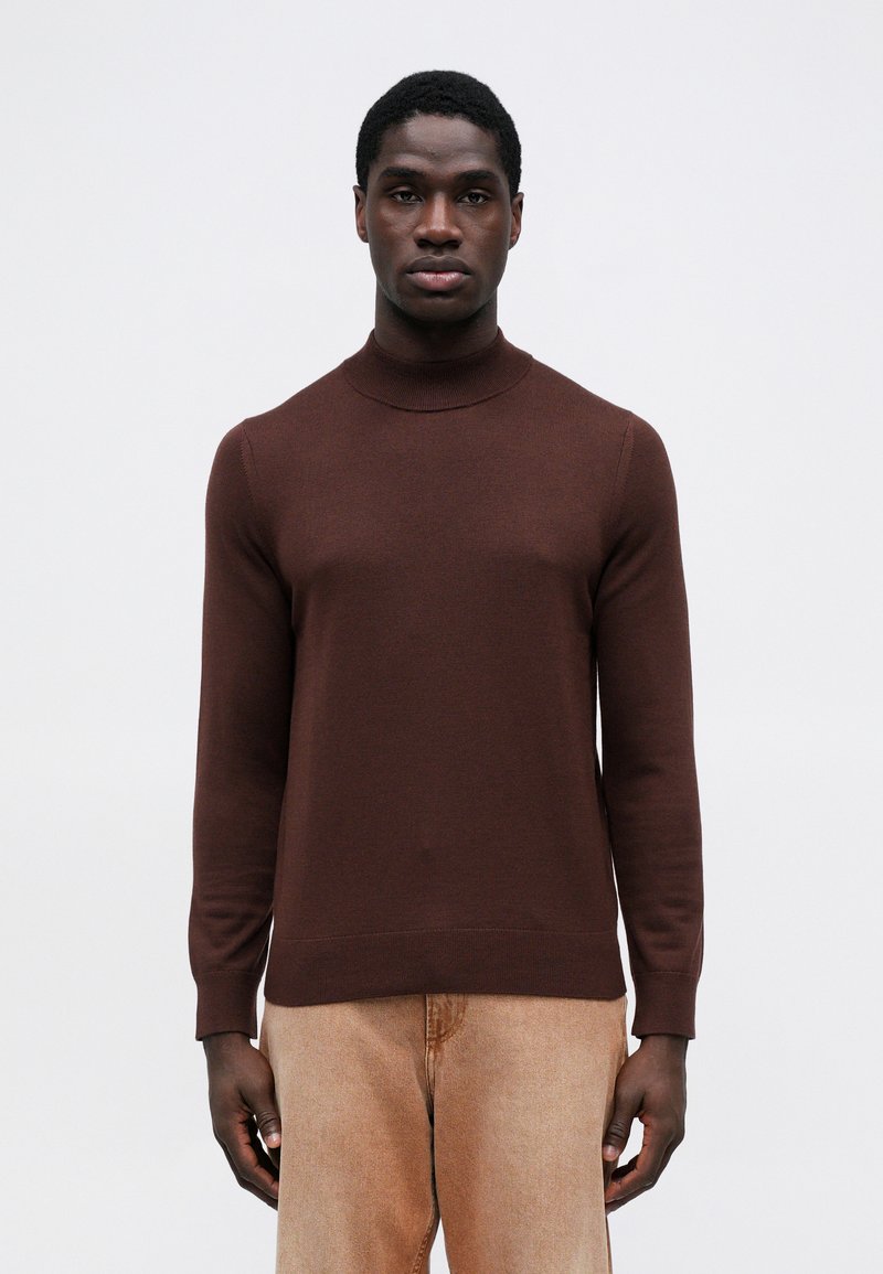 Brun, tætsiddende turtleneck sweater med lange ærmer, lavet af glat strikmateriale. Matchet med lysebeige bukser for kontrast.
