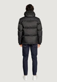 Giacca nera imbottita con colletto alto, caratterizzata da un accento del logo, indossata sopra jeans blu e scarpe da ginnastica bianche. Texture liscia e lucida.