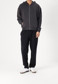 Grijze rits-hoodie met een voorzak, gecombineerd met zwarte loszittende broek en witte schoenen. Soepele stofstructuur en casual ontwerp.