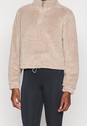 Pull beige duveteux avec demi-fermeture éclair et ourlet à cordon, porté avec des leggings noirs ajustés sur une personne debout devant un fond uni.