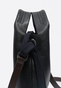 Čierna kožená taška z bočného pohľadu s tmavomodrým ťažkým pásom a hnedou koženou rukoväťou na bielom pozadí.