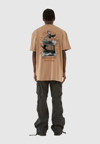 Tan oversized t-skjorte med trykk av et fuglebur og flyvende fugler på ryggen, kombinert med svarte cargo-bukser og en avslappet passform.