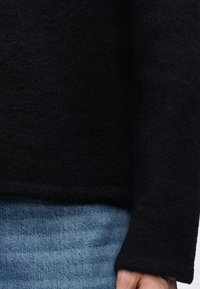 Pull noir texturé à manches longues porté avec un jean en denim bleu et la main droite visible sur le côté.