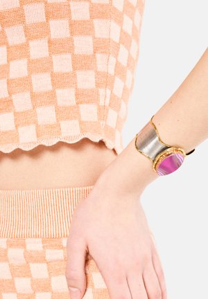 Bracciale in metallo dorato con una pietra centrale in agata rosa, dalle forme irregolari, combinato con una finitura in metallo argentato. Indossato su una mano.