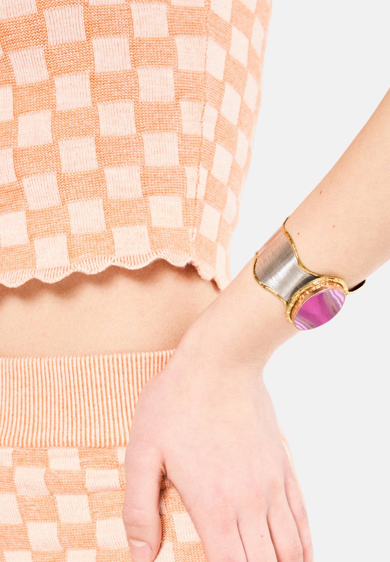 Guldfarvet metalarmbånd med en central pink agatsten, prydet med uregelmæssige former, kombineret med en sølvfarvet metalindlæg. Båret på en hånd.