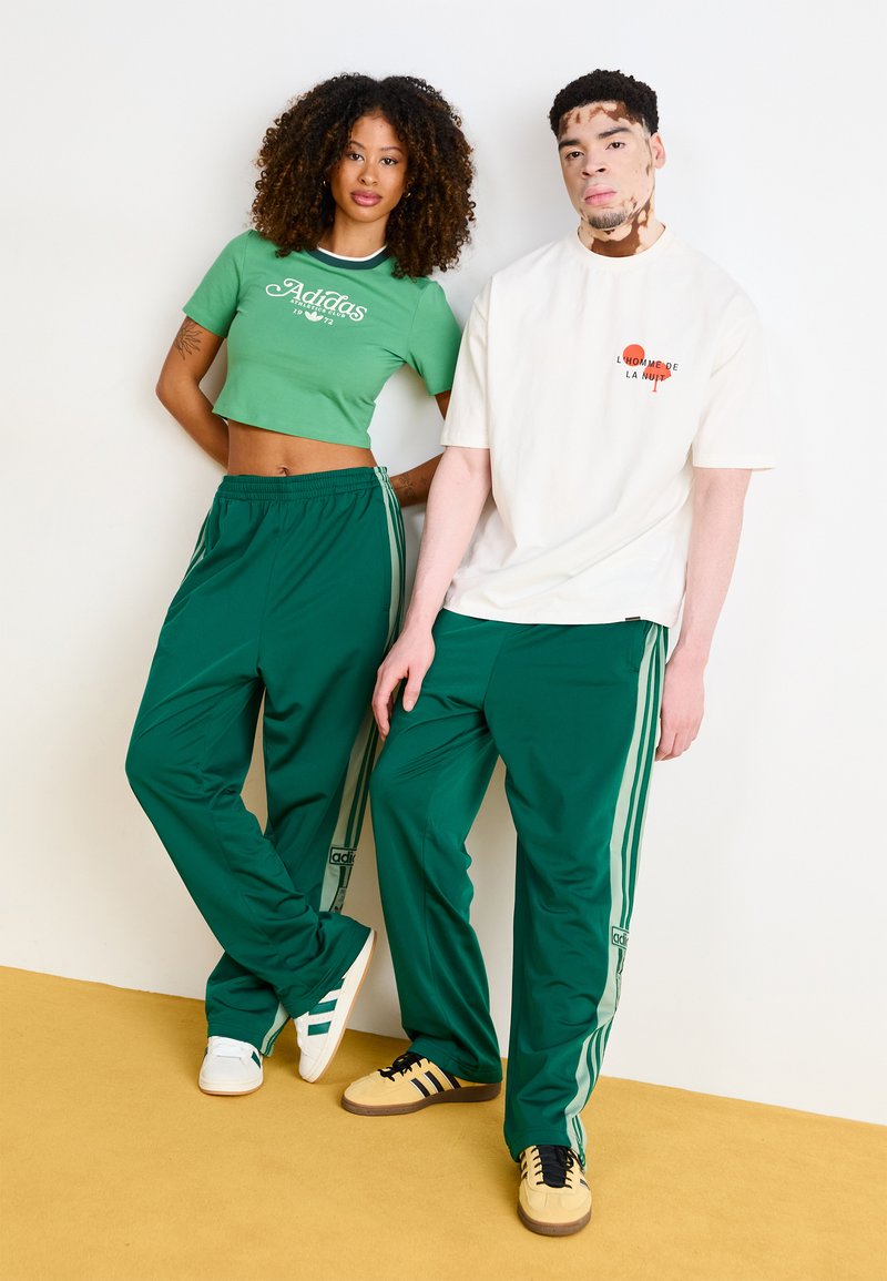 Top corto verde y pantalones deportivos verdes a juego con rayas laterales. camiseta blanca con texto rojo; hombre lleva zapatos negros y marrones, mujer zapatos blancos y verde azulado.