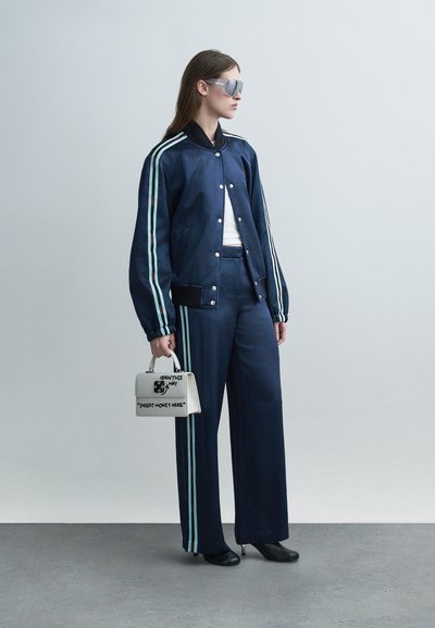 Γυναίκα με μπλε σκούρο αθλητικό σετ με λευκές ρίγες, λευκό τοπ, μαύρες γόβες, ανακλαστικά γυαλιά ηλίου, κρατώντας λευκή τσάντα χειρός με μαύρο κείμενο.