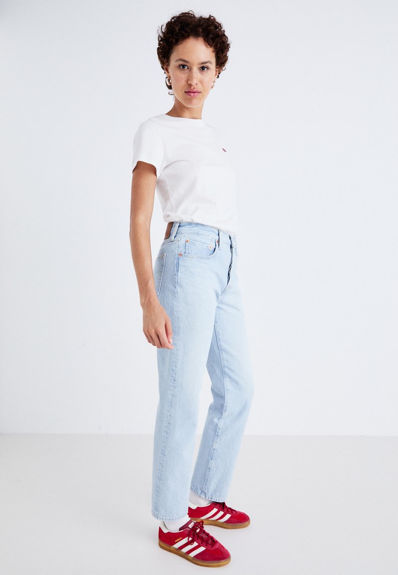 Maglietta bianca a maniche corte con un piccolo logo rosso, jeans in denim chiaro a vita alta, e sneakers rosse con strisce bianche sui lati.