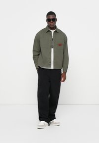 Green button-up jacket with two chest pockets, worn over a white t-shirt, paired with black cargo pants and light-colored sneakers.
