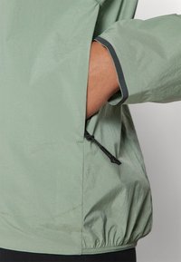Helle grüne, leichte Jacke aus strukturiertem Stoff, mit elastischem Saum und einer zipbaren Tasche mit schwarzem Zugband und Kontrastnähten.