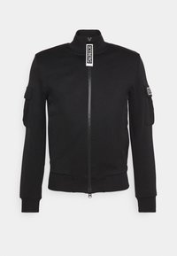 Chaqueta negra con cremallera hecha de tela suave, que presenta un cuello alto, puños de canalé y bolsillos laterales. Incluye un parche con el logo en la manga.