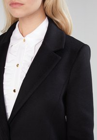 Sort uld blazer med en struktureret krave, der har en hvid flæsebluse under. Guldknapdetaljer på blusen er synlige.