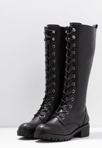 Bottes noires en cuir montant jusqu'aux genoux, avec un bout pointu, une semelle plate et un laçage à l'avant avec des œillets métalliques et une surface texturée.