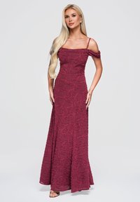 Femme blonde portant une robe de soirée bordeaux sans manches et épaules dénudées avec des motifs circulaires texturés, debout devant un fond uni.